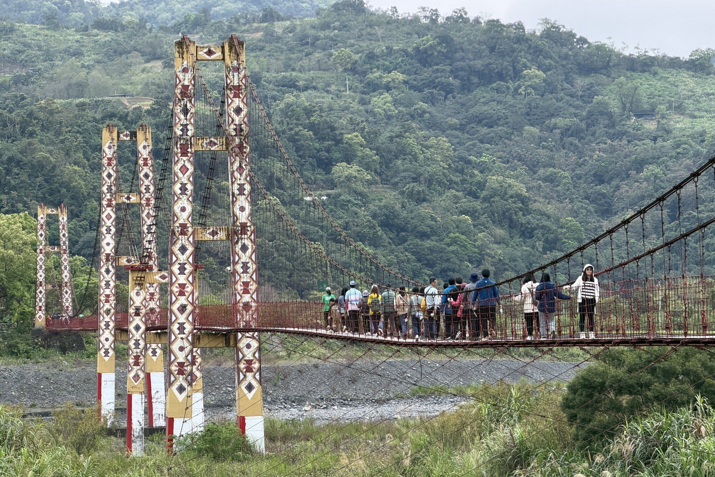 一群人一起走過繪有原住民族傳統紋樣的吊橋，背景是山林與河谷