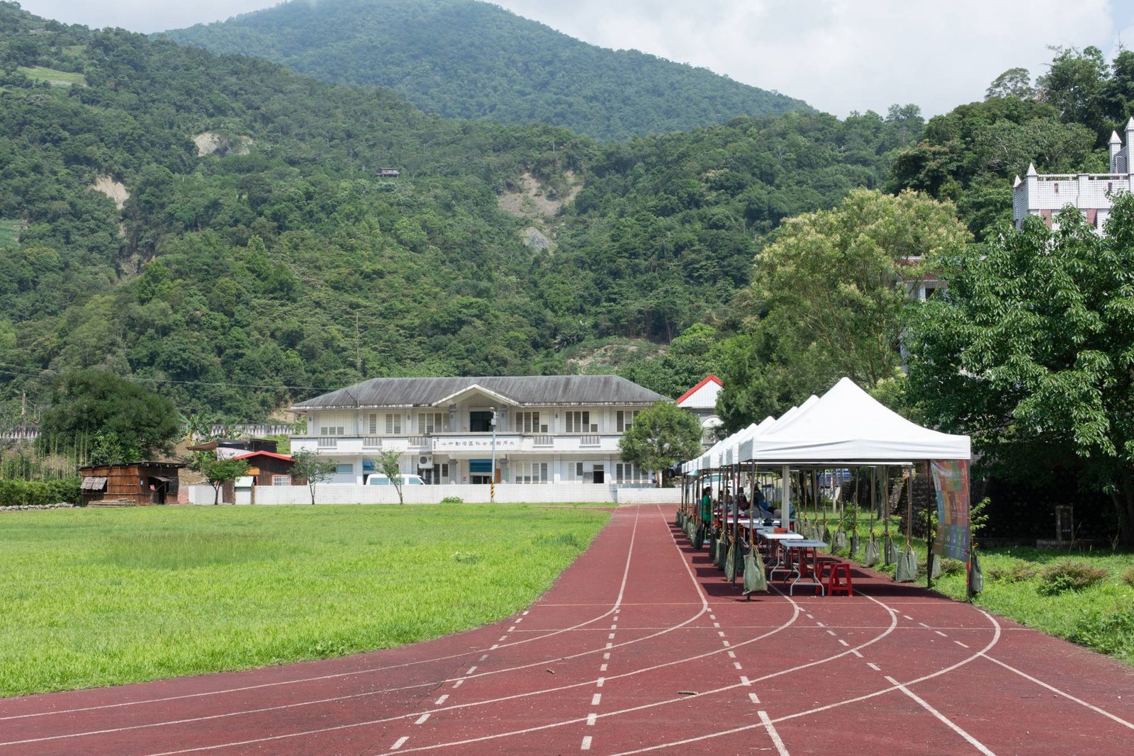 White tents and registration tables set up beside a running track, with a community center and forested hills behind