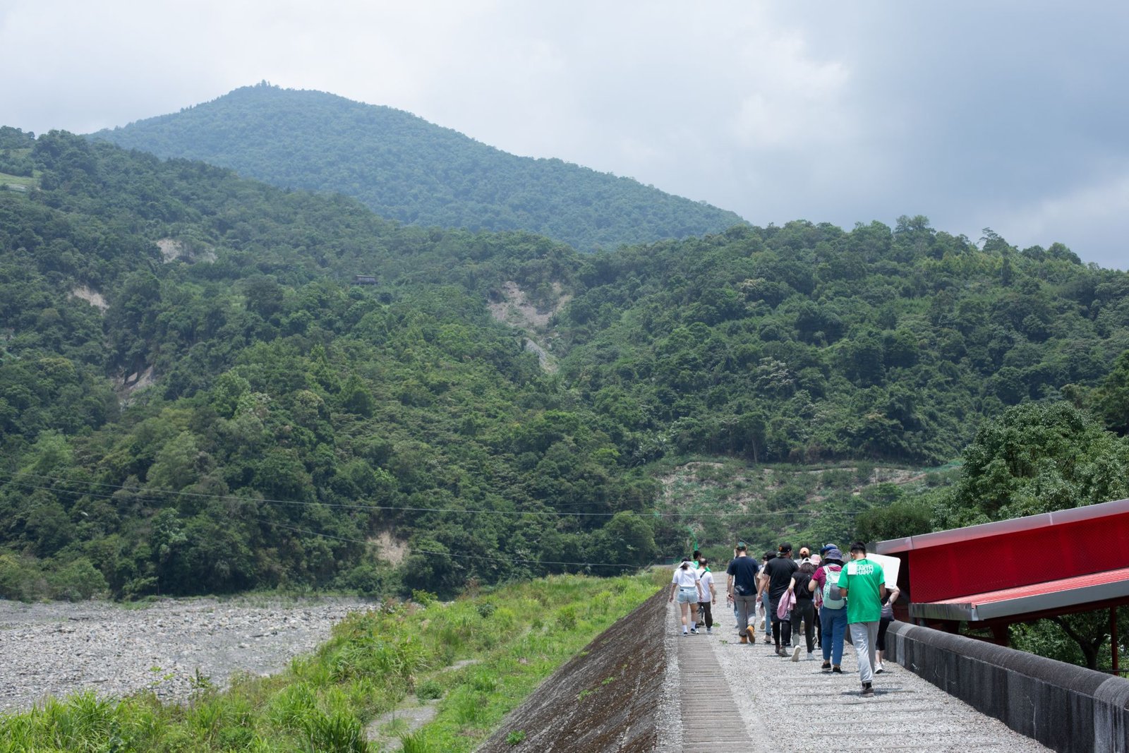 A group of people walking along a riverside path toward forested mountains, one wearing a green DOMI EARTH HAPPY shirt